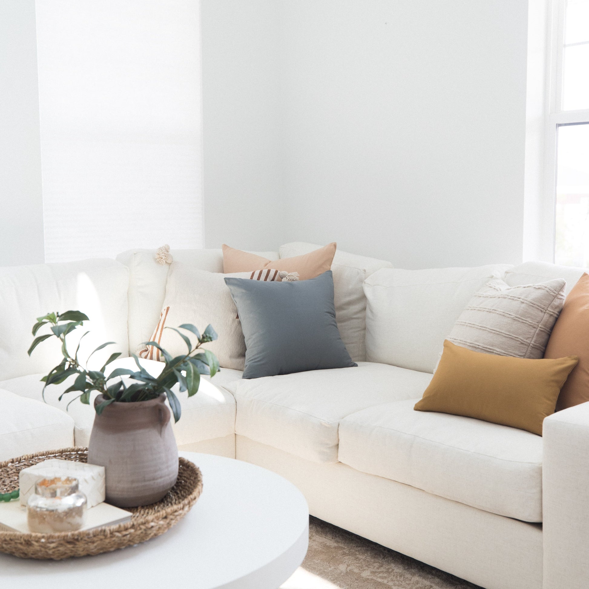 Cream sectional sofa styled with gray, tan, and striped throw pillows in a bright room with potted plant and round table.