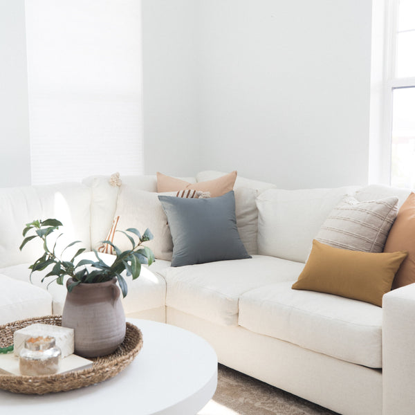 Cream sectional sofa styled with gray, tan, and striped throw pillows in a bright room with potted plant and round table.