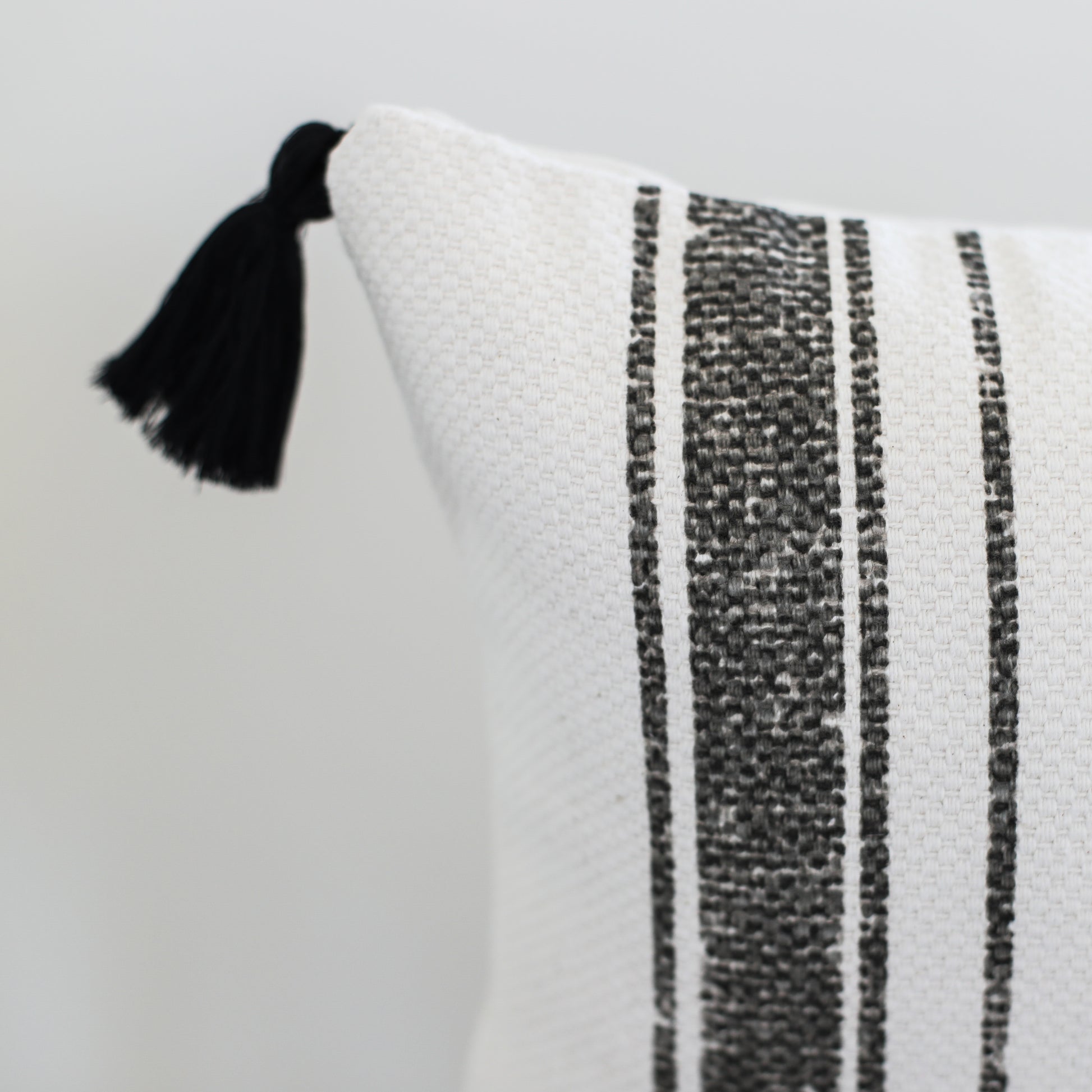 White lumbar pillow with charcoal striped pattern and black tassel corner detail.
