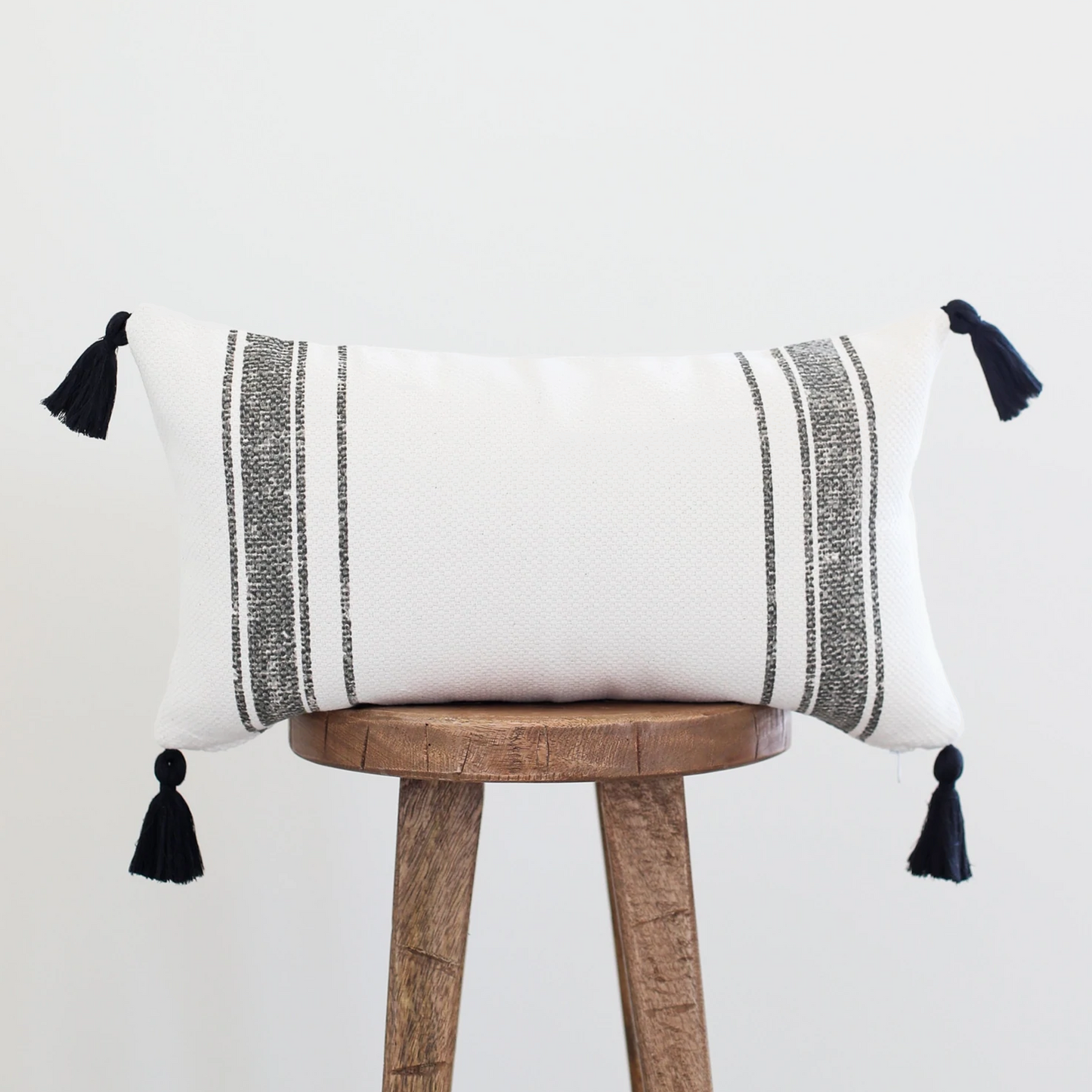 White lumbar pillow with gray striped pattern and black corner tassels on wooden stool.
