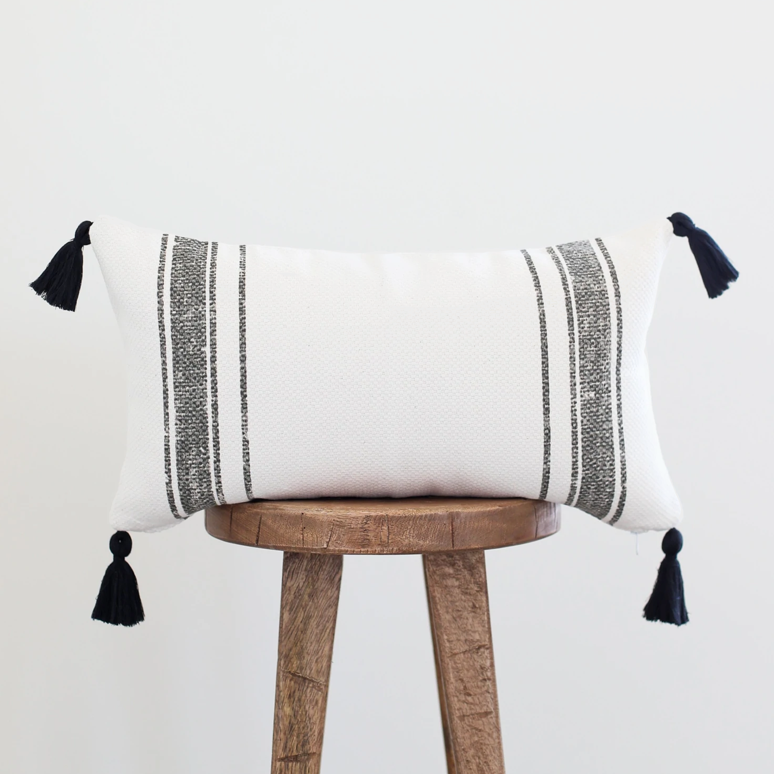 White lumbar pillow with gray striped pattern and black corner tassels on wooden stool.