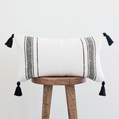 White lumbar pillow with gray striped pattern and black corner tassels on wooden stool.