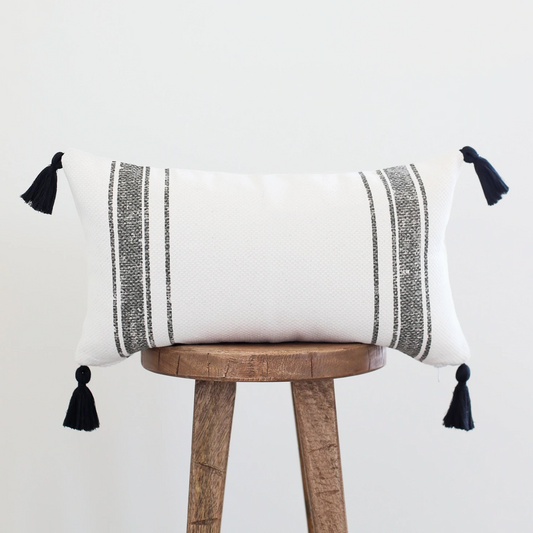 White lumbar pillow with gray striped pattern and black corner tassels on wooden stool.
