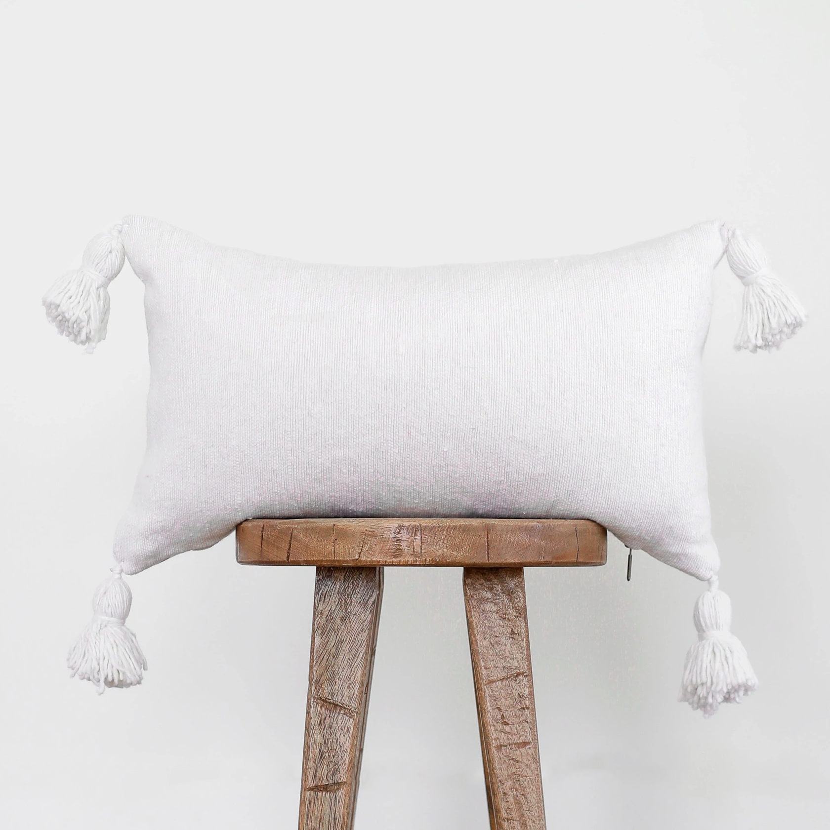 White lumbar pillow with tassel corners displayed on wooden stool.
