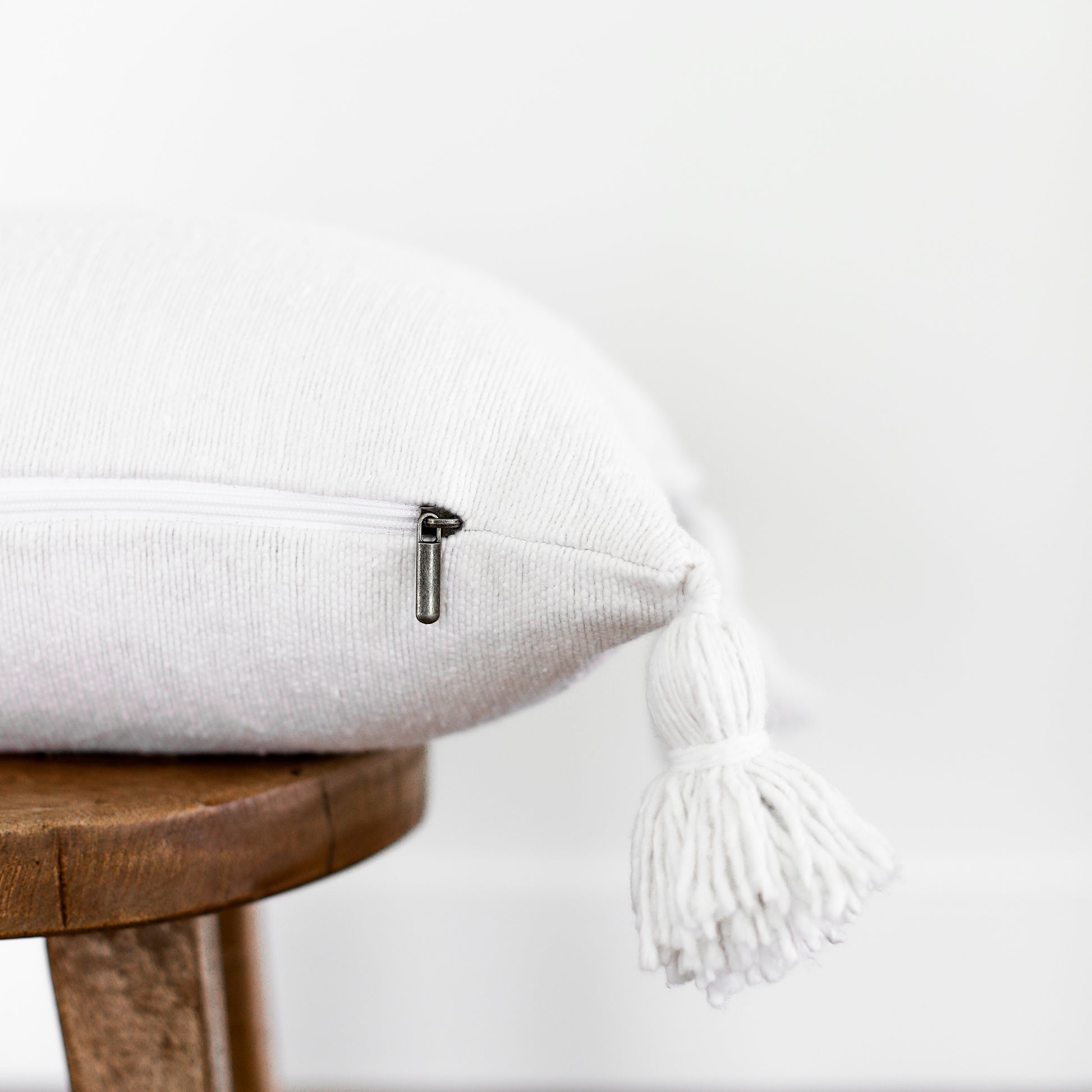 White lumbar pillow with textured fabric and white tassels on corners, displayed on wooden stool.