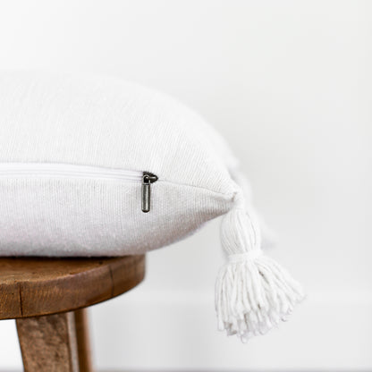 White lumbar pillow with textured fabric and white tassels on corners, displayed on wooden stool.
