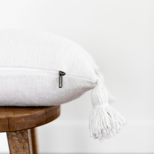 White lumbar pillow with textured fabric and white tassels on corners, displayed on wooden stool.