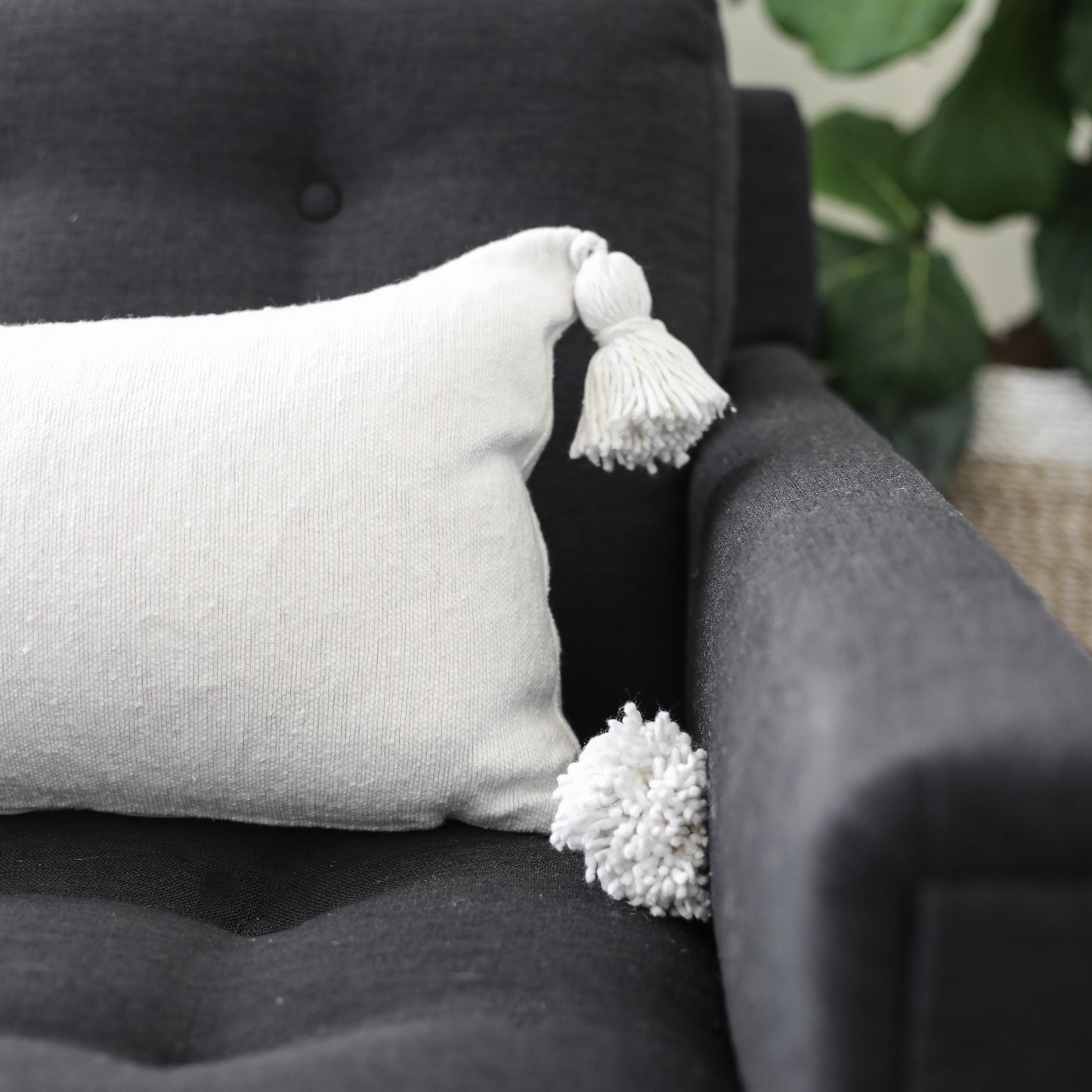 Cream lumbar pillow with white tassels on corners, displayed on dark gray upholstered furniture.