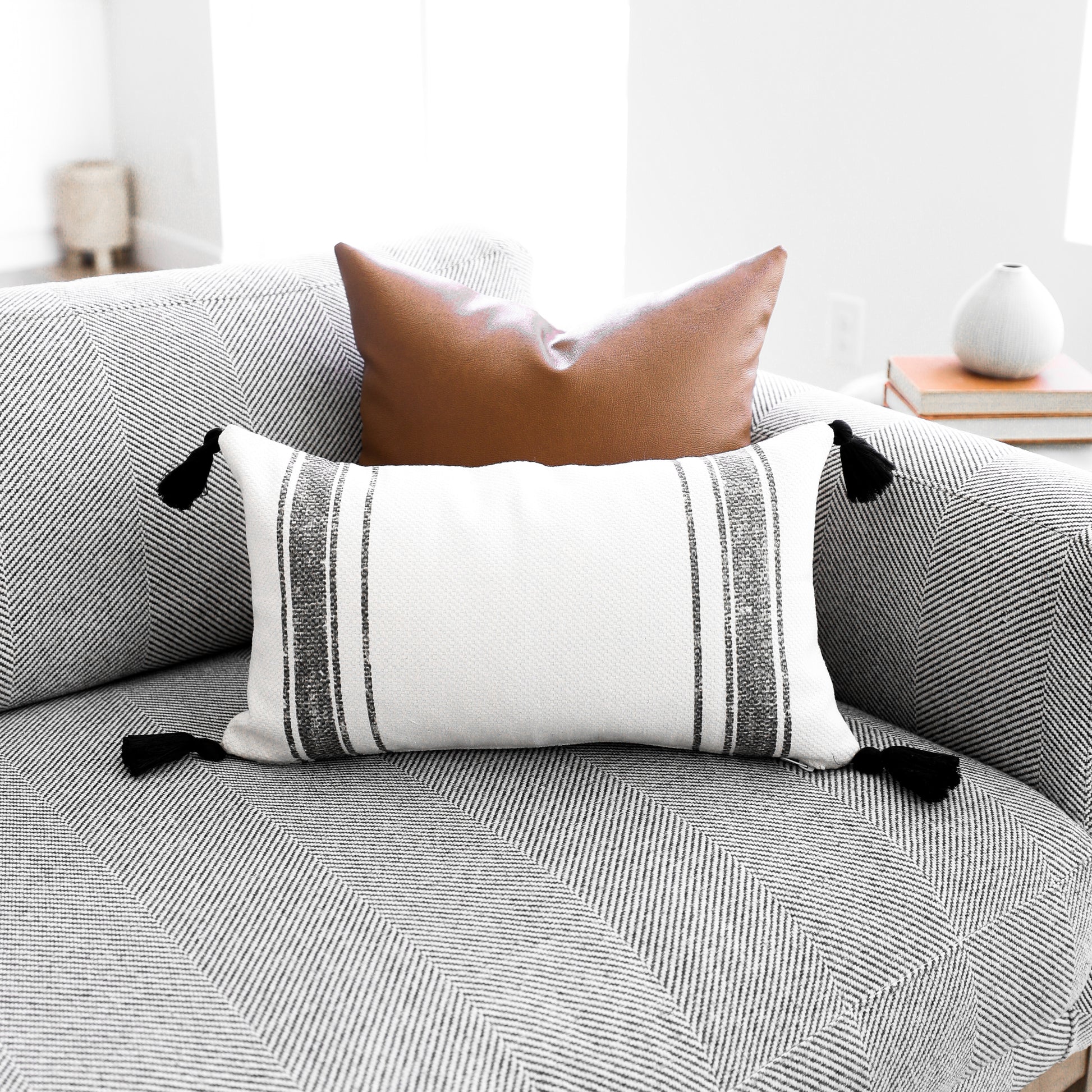 Rectangular lumbar pillow with cream and gray striped pattern and black tassel corners, displayed on a gray upholstered sofa.