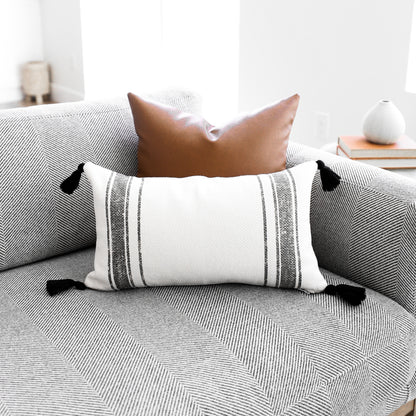 Rectangular lumbar pillow with cream and gray striped pattern and black tassel corners, displayed on a gray upholstered sofa.