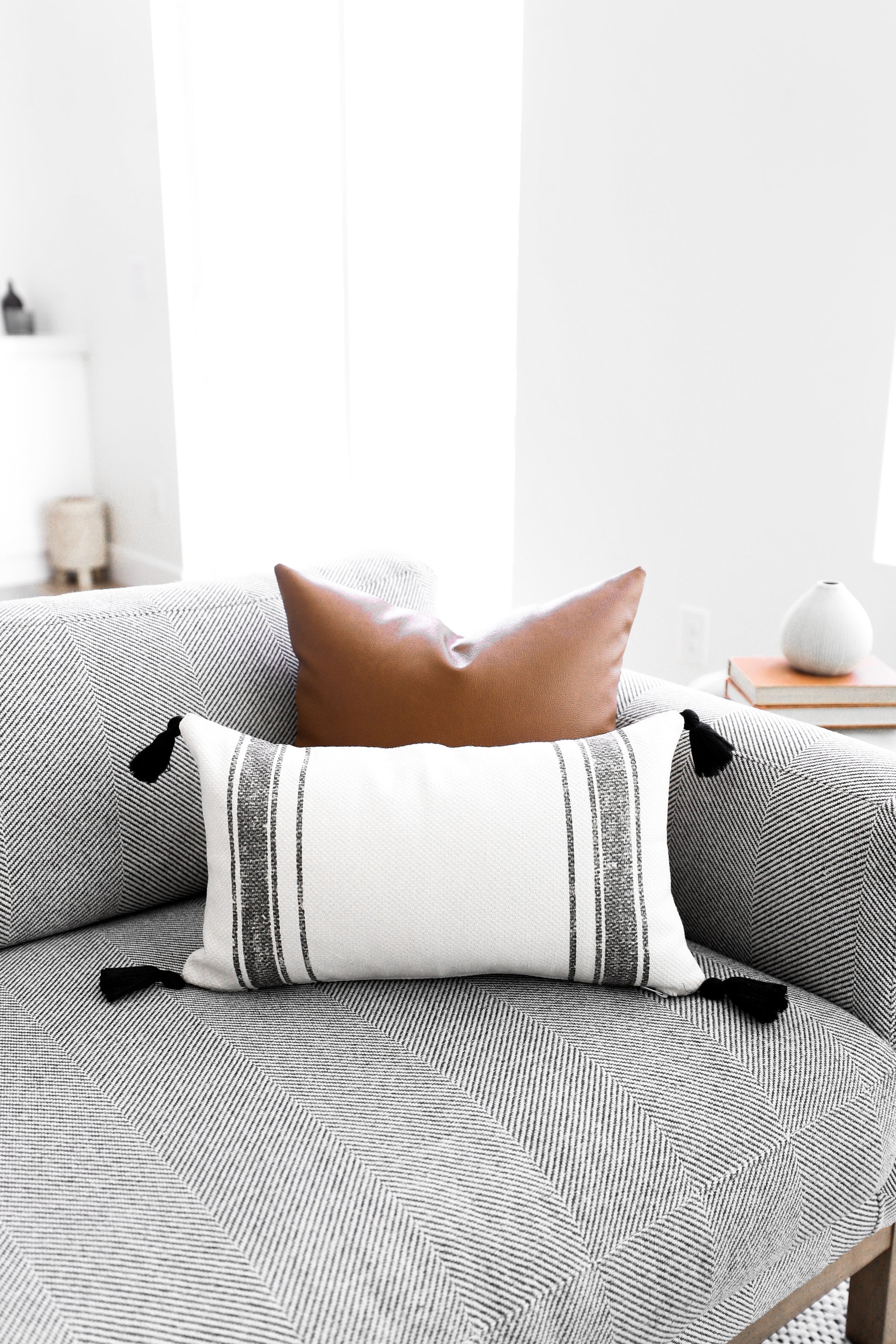 White lumbar pillow with gray horizontal stripes and black corner tassels, displayed on a gray geometric-patterned sofa.