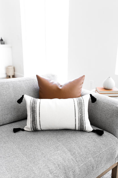 White lumbar pillow with gray horizontal stripes and black corner tassels, displayed on a gray geometric-patterned sofa.