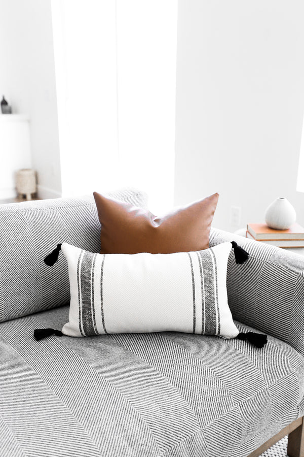 White lumbar pillow with gray horizontal stripes and black corner tassels, displayed on a gray geometric-patterned sofa.