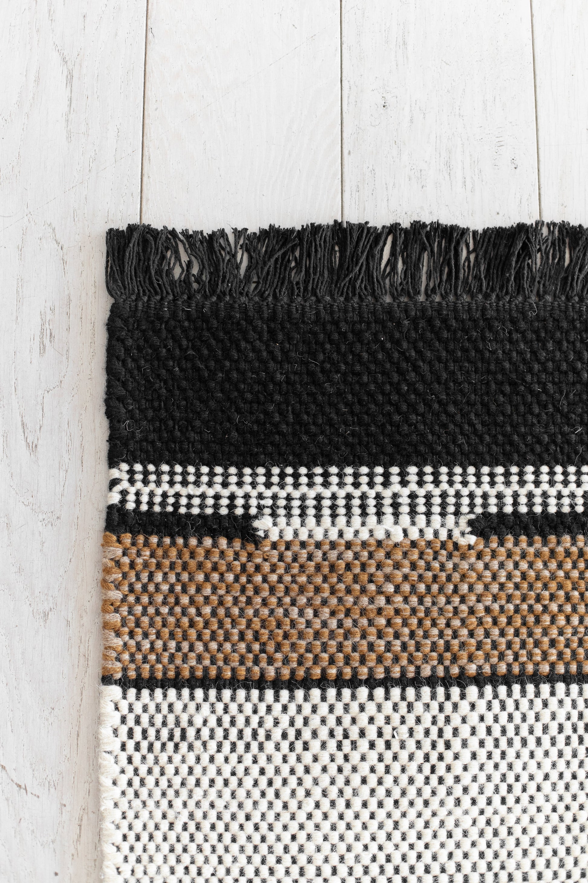 Black, white, and tan striped woven textile with fringe trim on white painted wood surface.