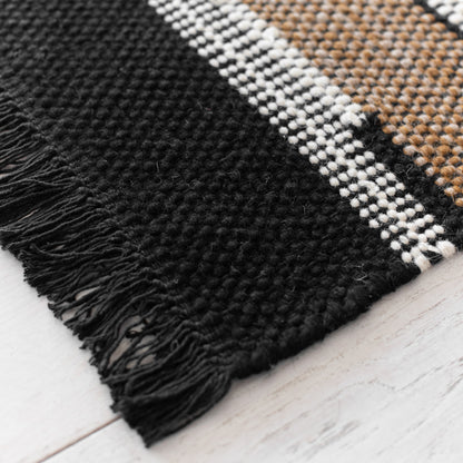 Woven rug with black, white, and tan geometric stripes with black fringe edges on light wood floor.