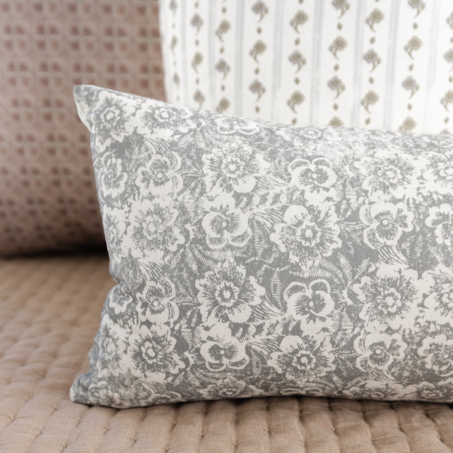 Gray and white lumbar pillow with ornate floral damask pattern, displayed on wooden floor with patterned fabrics in backgroun