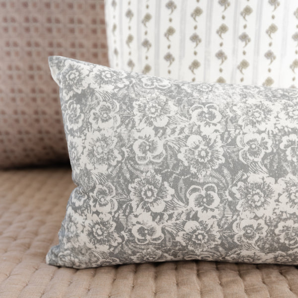 Gray and white lumbar pillow with ornate floral damask pattern, displayed on wooden floor with patterned fabrics in backgroun