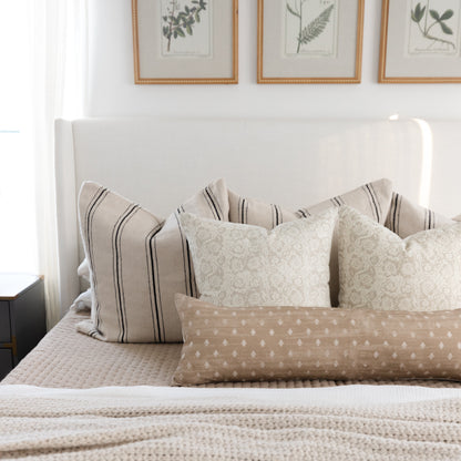 Tan lumbar pillow with white dotted pattern displayed on bed with striped and patterned pillows.