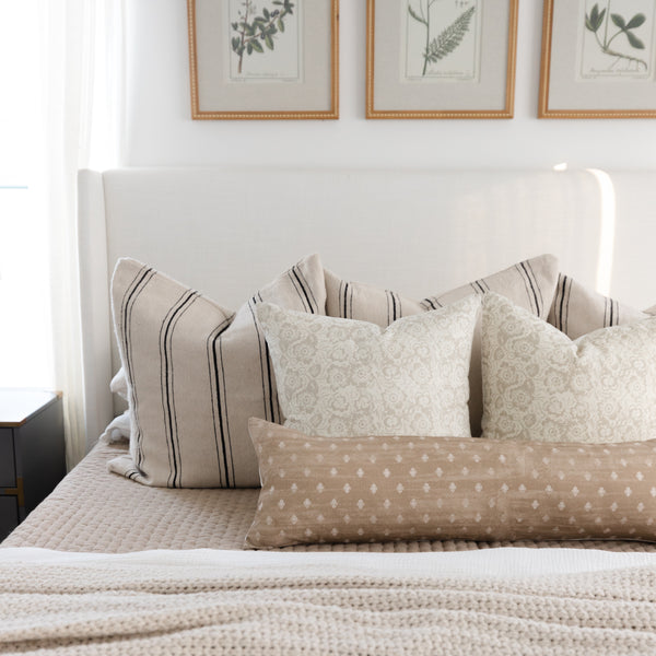 Tan lumbar pillow with white dotted pattern displayed on bed with striped and patterned pillows.