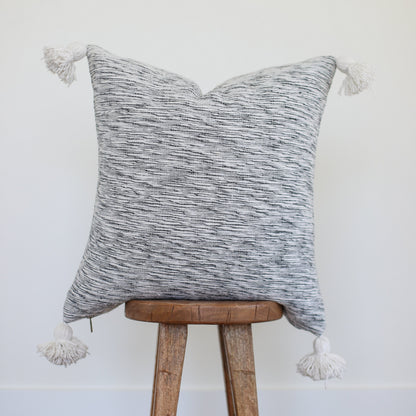 Gray and white textured throw pillow with tassel corners on a wooden stool.