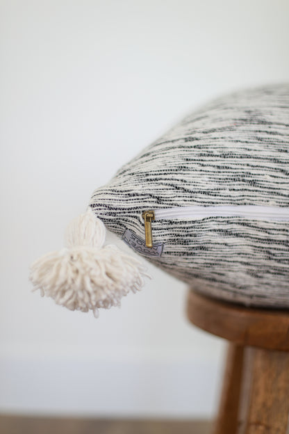 Round pillow with black and white striped woven fabric, cream fringe trim, and wooden base with brass hardware detail.