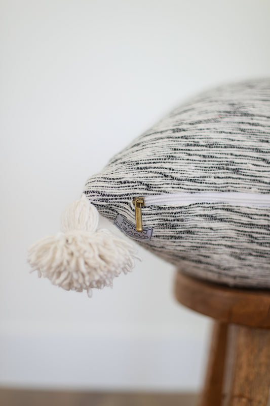 Round pillow with black and white striped woven fabric, cream fringe trim, and wooden base with brass hardware detail.