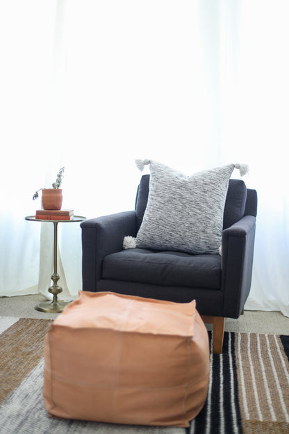 Dark upholstered armchair with gray textured pillow, tan pouf, side table with potted plant, and striped rug in room setting.
