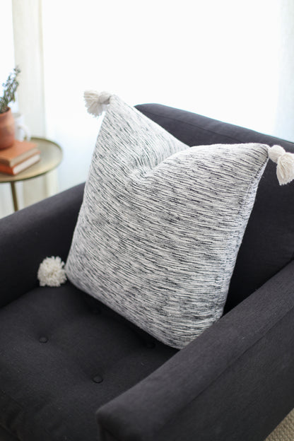 Gray and white textured throw pillow with white tassel corners on black sofa.