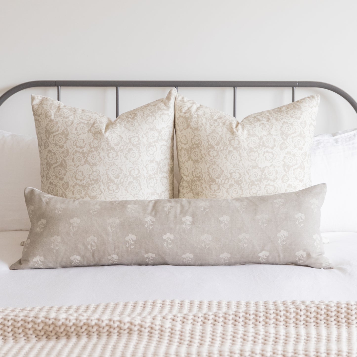 Three beige pillow covers with floral damask and botanical patterns arranged on a bed with metal headboard.