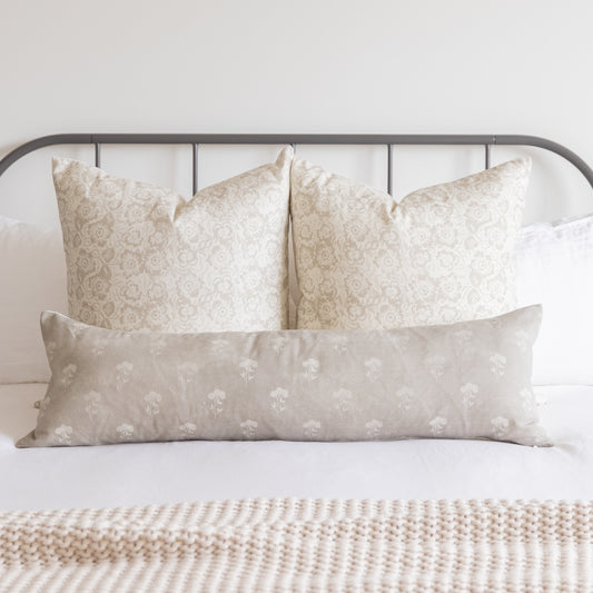Three beige pillow covers with floral damask and botanical patterns arranged on a bed with metal headboard.