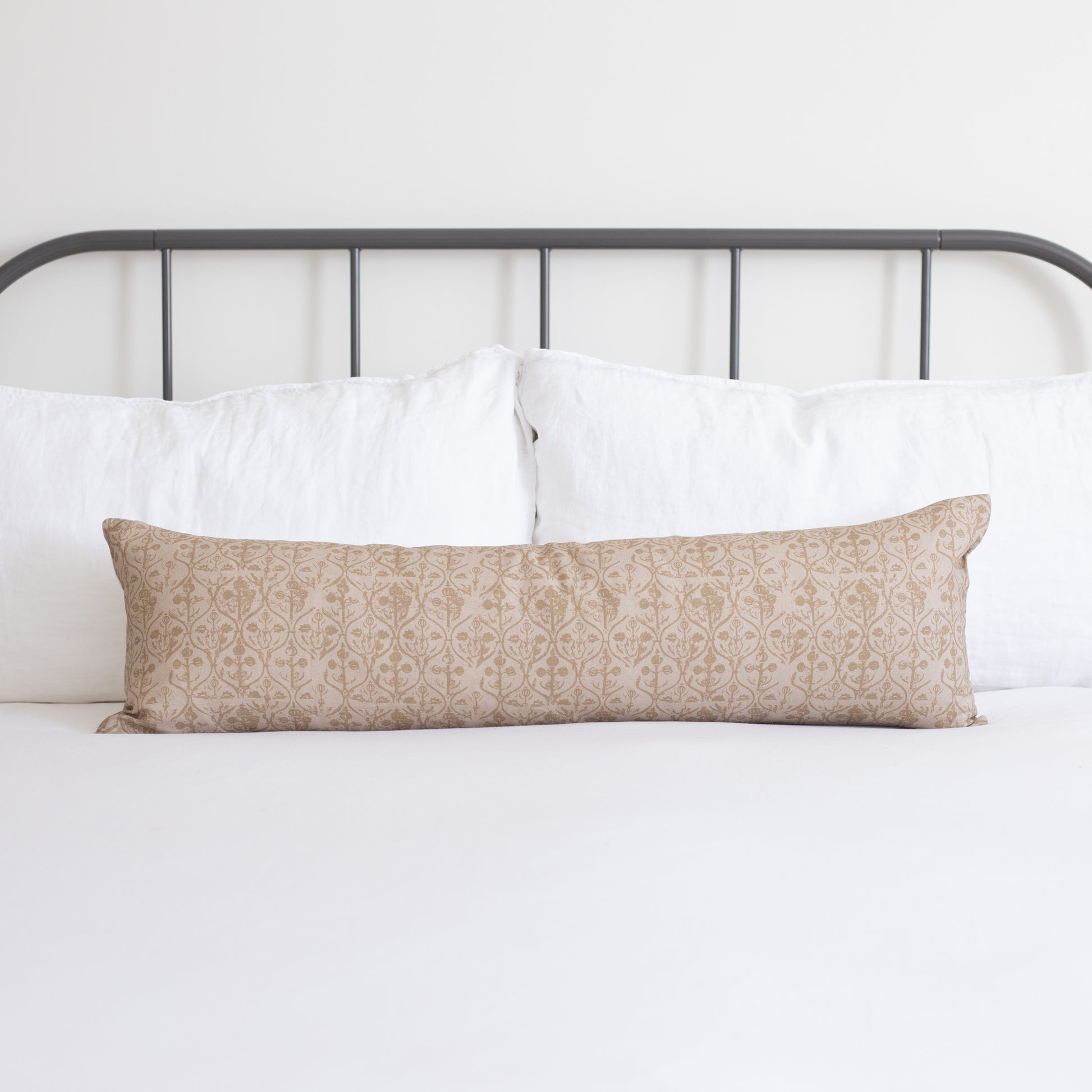 Tan lumbar pillow with small-scale geometric floral pattern displayed on bed with white pillows and metal headboard.