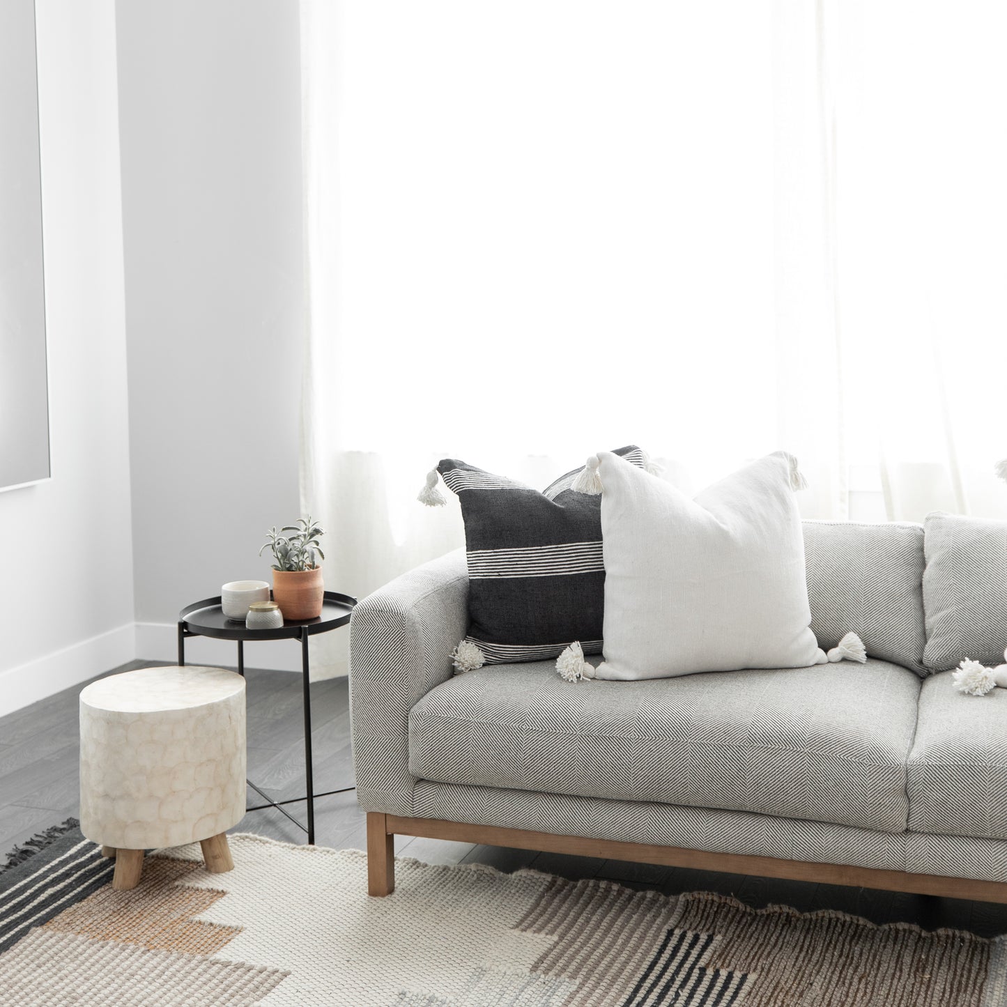 White square throw pillow with tassels displayed on a gray sofa beside black and striped accent pillows.