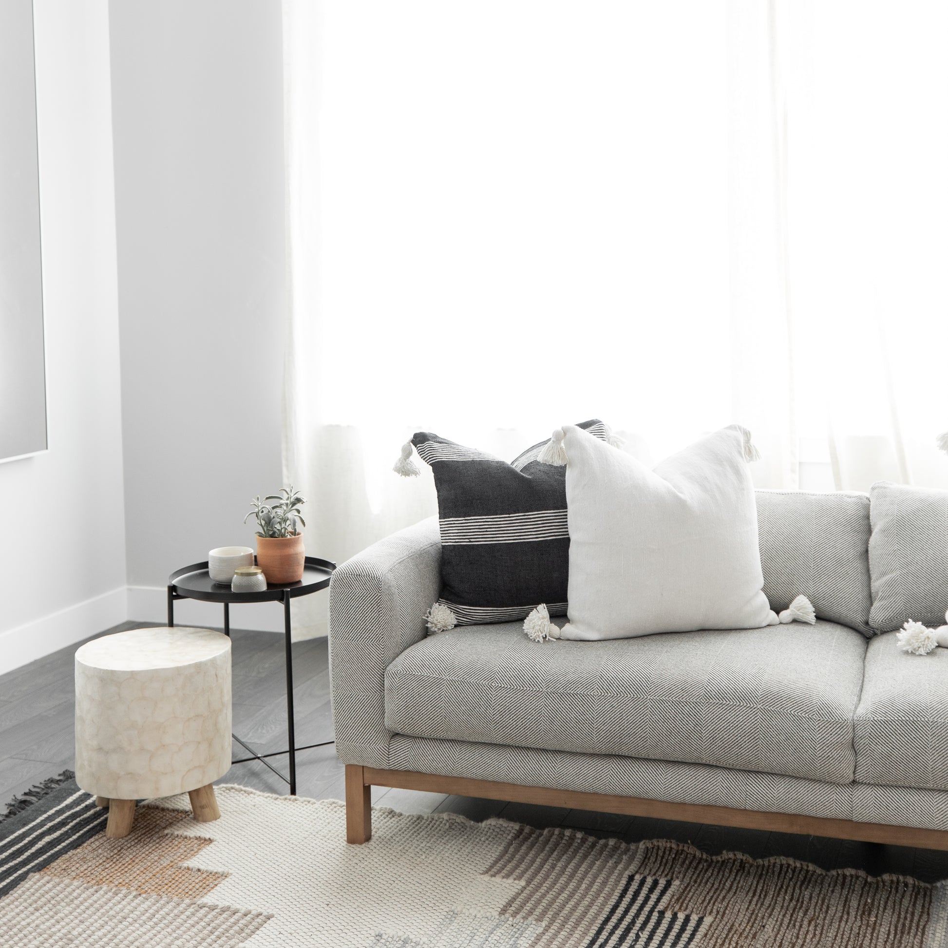 White square throw pillow with tassels displayed on a gray sofa beside black and striped accent pillows.