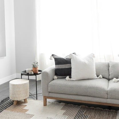 White square throw pillow with tassels displayed on a gray sofa beside black and striped accent pillows.