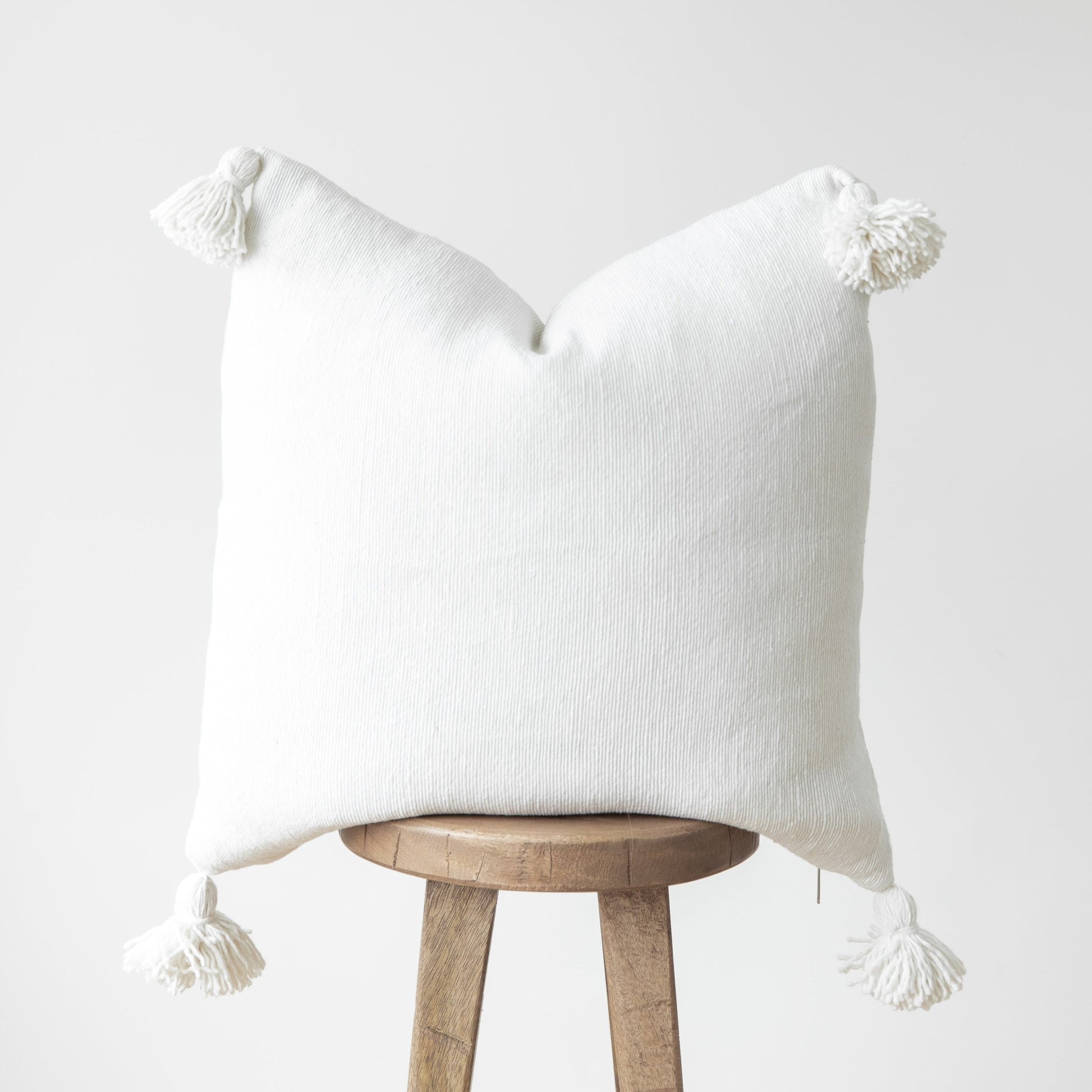 White textured throw pillow with corner tassels draped over a wooden stool.
