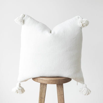 White textured throw pillow with corner tassels draped over a wooden stool.