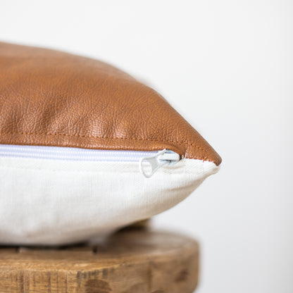 Two-tone rectangular pillow with tan leather top and cream linen bottom, zipper closure visible, resting on wooden base.