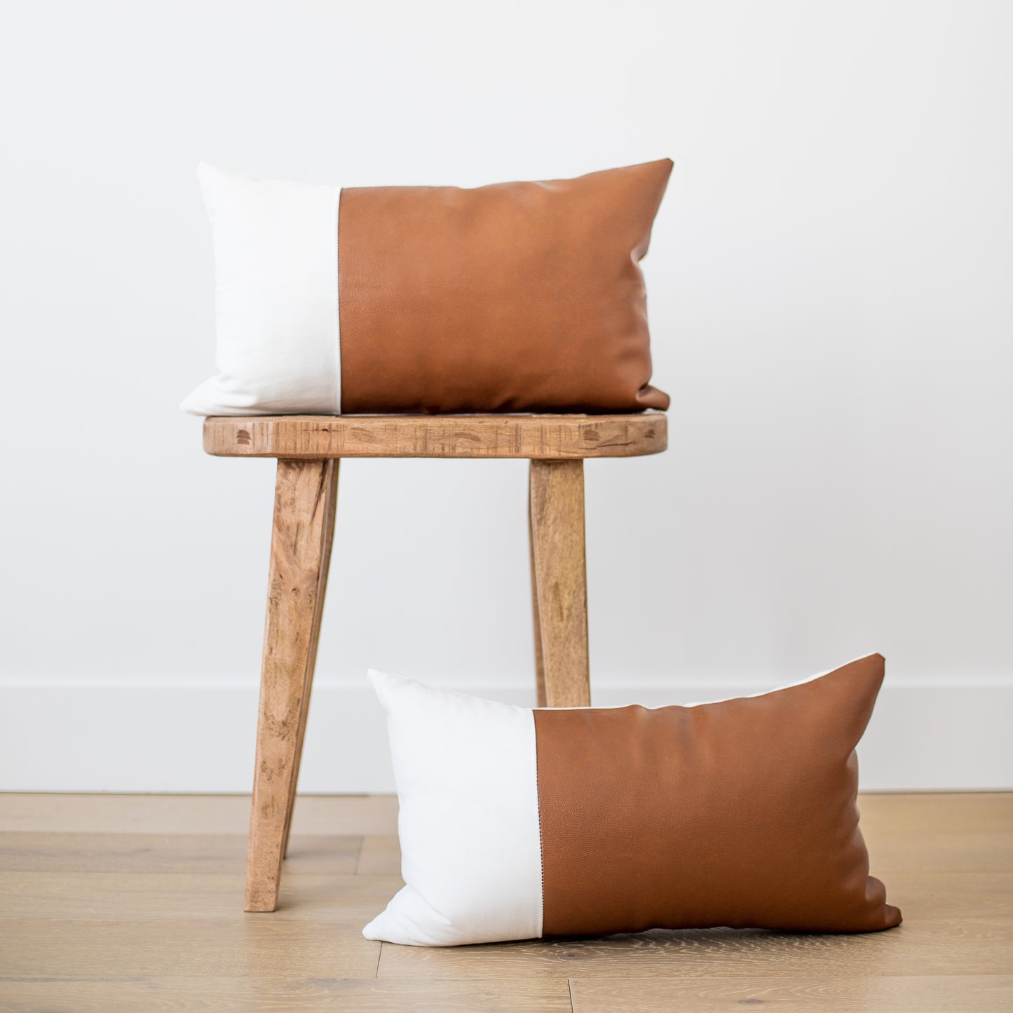 Two rectangular pillows with tan leather and white linen colorblock design displayed on wooden stool and floor.