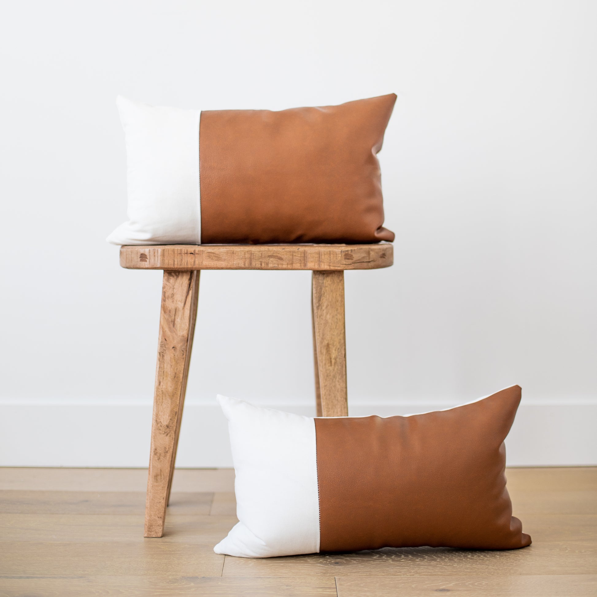 Two rectangular pillows with tan leather and white linen colorblock design displayed on wooden stool and floor.
