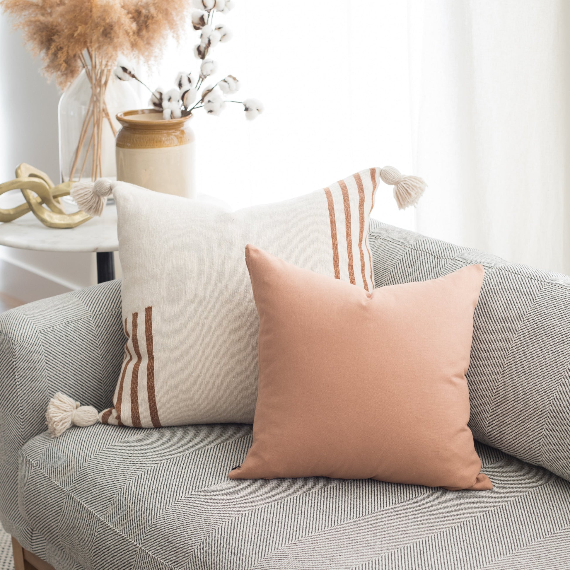 Two decorative pillows on a gray textured sofa: cream with rust stripes and tassels, and solid blush pink.