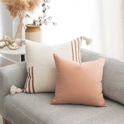 Two decorative pillows on a gray textured sofa: cream with rust stripes and tassels, and solid blush pink.