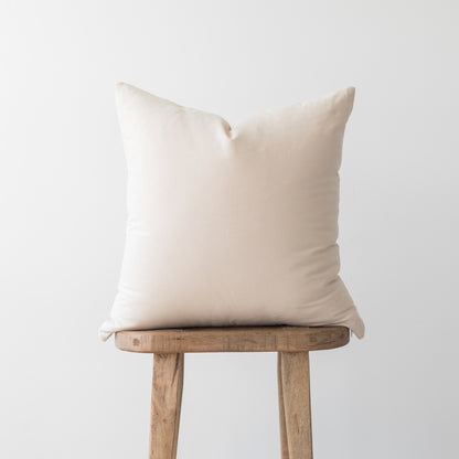 Cream-colored throw pillow with square shape displayed on wooden stool.
