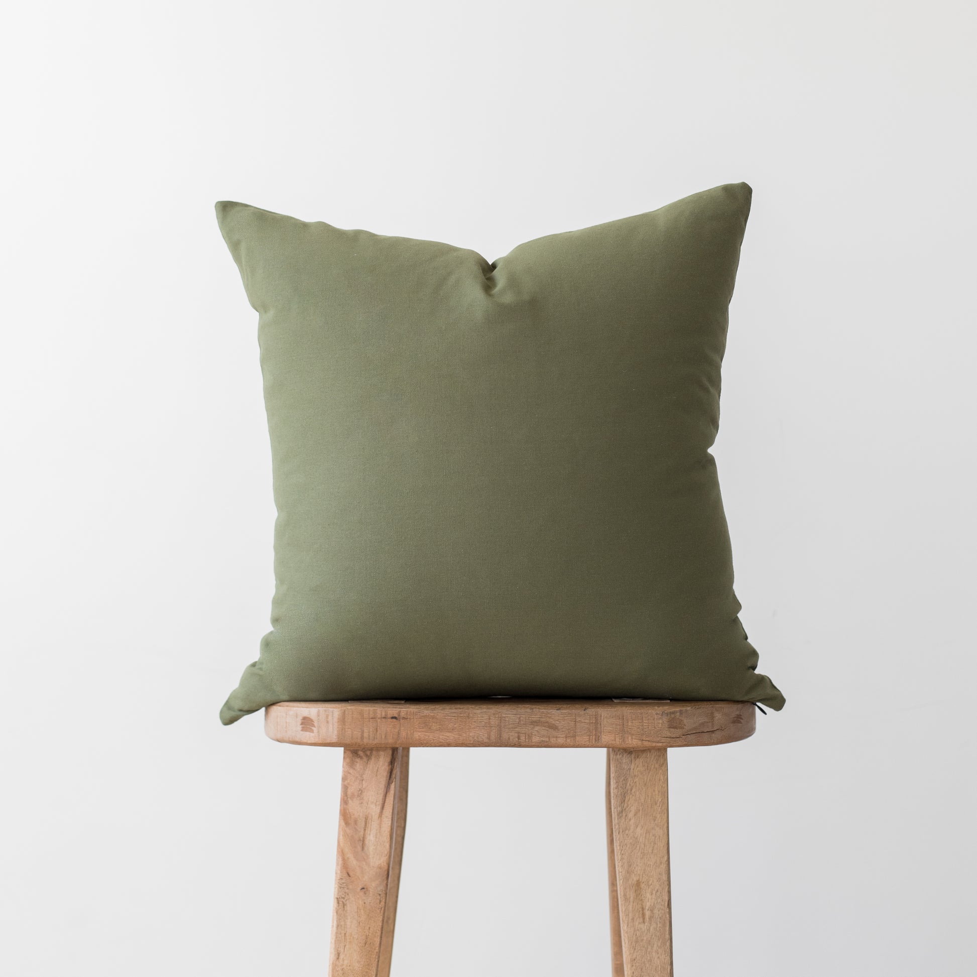 Olive-colored square throw pillow with envelope closure displayed on wooden stool.