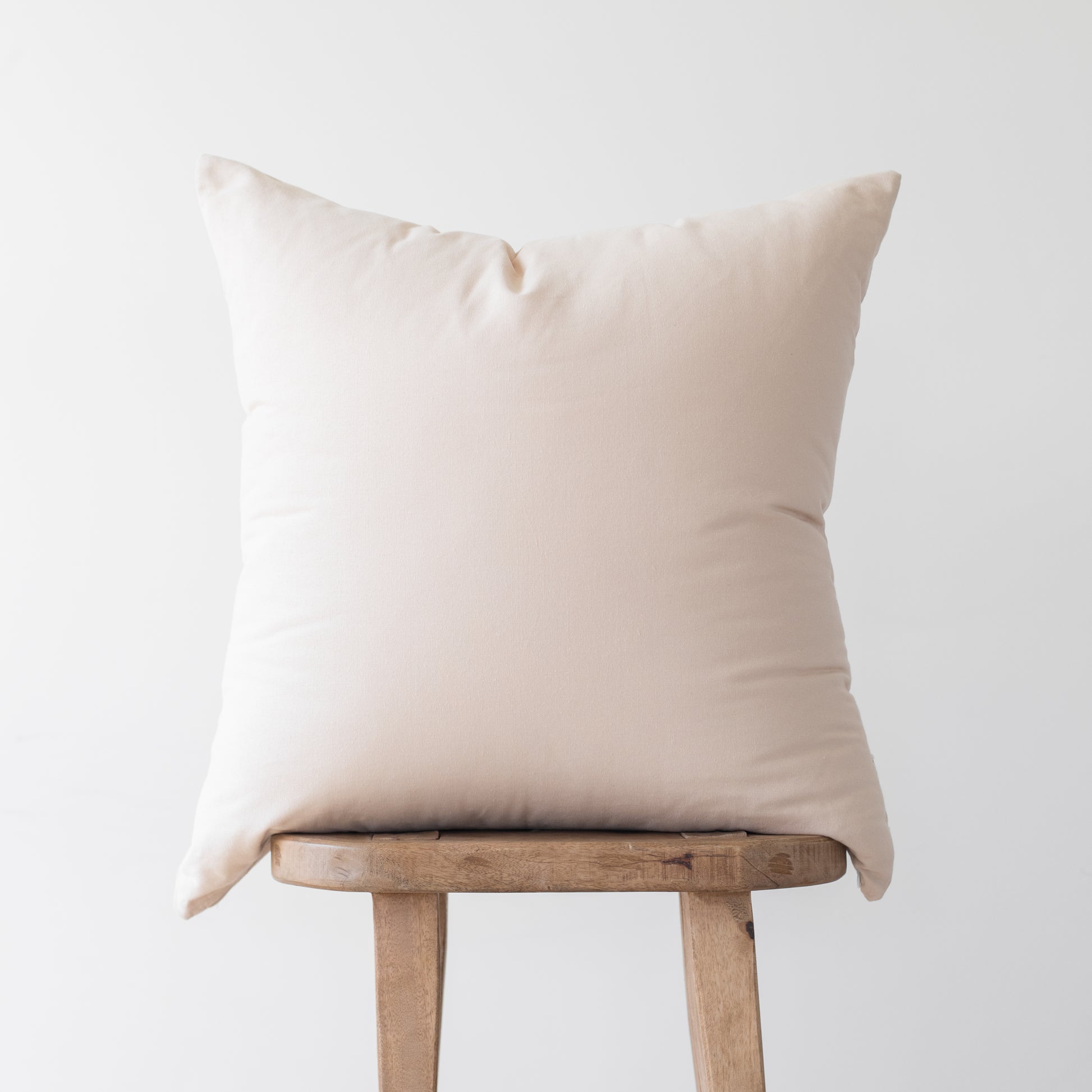 Cream-colored throw pillow with envelope closure displayed on wooden chair.