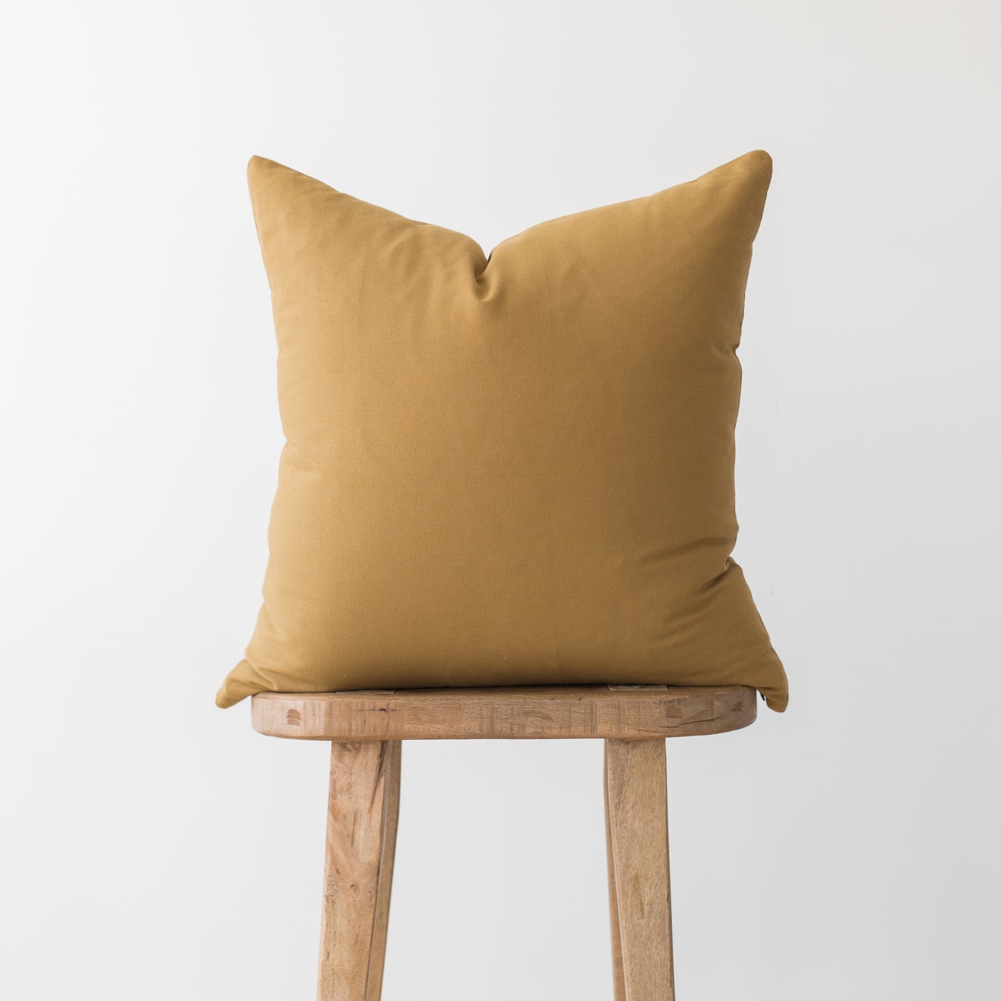 Solid gold-toned cushion cover displayed on a wooden stool against a white background.