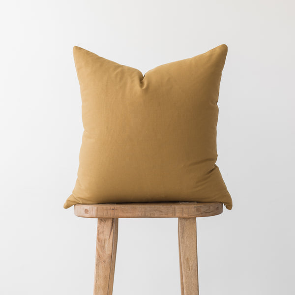 Solid gold-toned cushion cover displayed on a wooden stool against a white background.