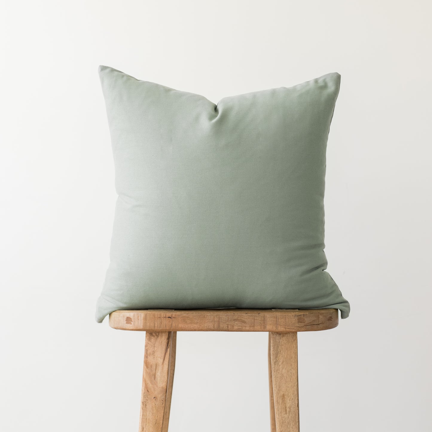 Sage green square pillow with solid linen texture displayed on wooden stool.