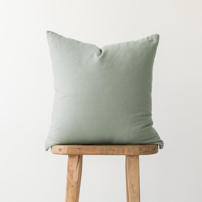 Sage green square pillow with solid linen texture displayed on wooden stool.