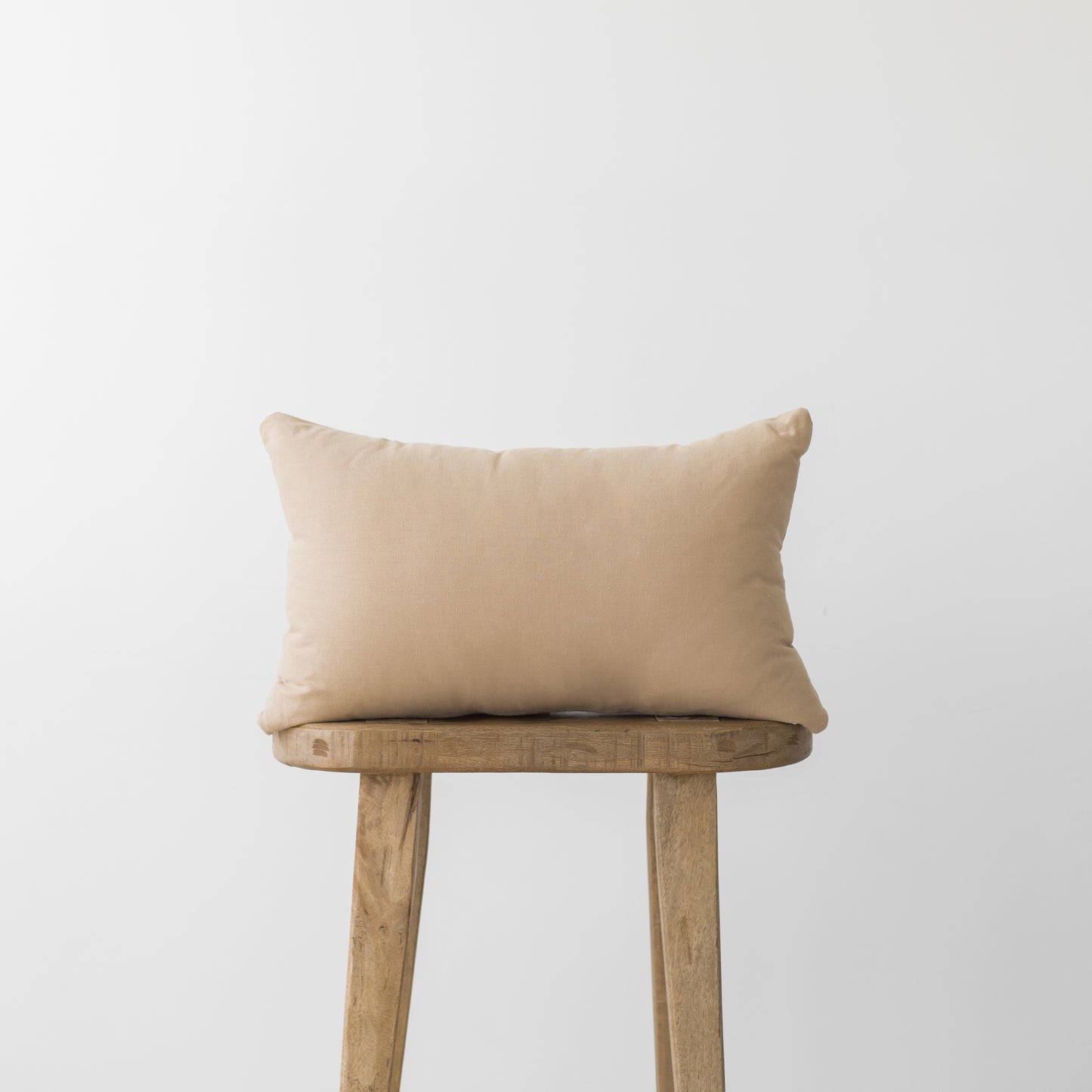 Beige rectangular pillow with corners resting on a wooden stool against a light gray background.