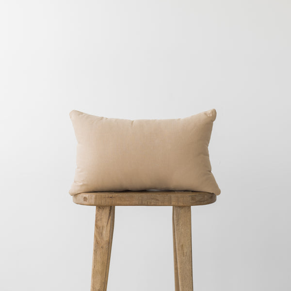 Beige rectangular pillow with corners resting on a wooden stool against a light gray background.